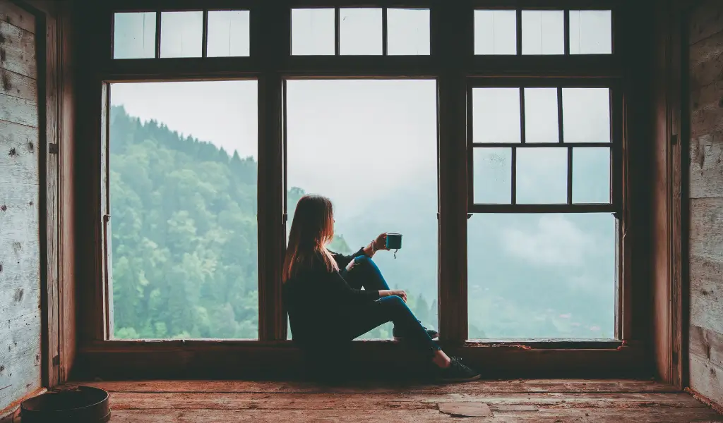 Kobieta siedzi z filiżanką herbaty, relaksując się przy oknie, wpatrując się spokojnie w krajobraz za szybą. Wokół niej panuje przytulna, domowa atmosfera w stylu Hygge.