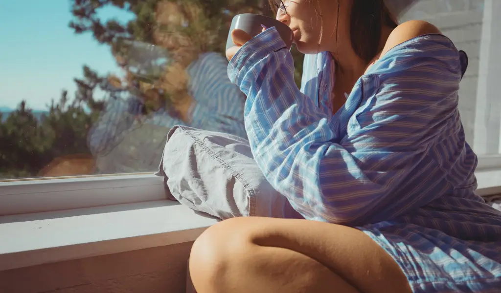 Dziewczyna z kubkiem kawy na parapecie patrzy przez okno – chwila spokoju i minimalizm w życiu.