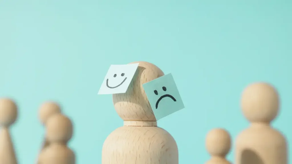 Zdjęcie ilustrujące 10 zasad zdrowia psychicznego – pionki z etykietami, z których jedne mają uśmiech, a inne ponurą minę, symbolizujące różne stany emocjonalne i kondycję psychiczną. W tle elementy nawiązujące do równowagi i spokoju, podkreślające znaczenie dbania o zdrowie psychiczne.