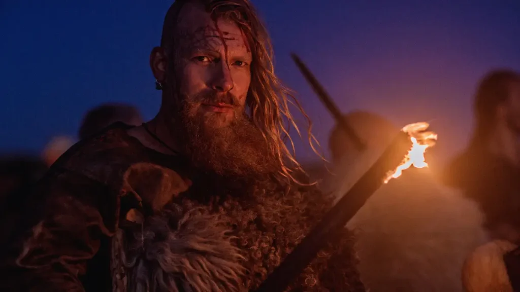 Wiking stojący nocą z pochodnią na tle fiordu, ubrany w tradycyjny strój wojownika z epoki wikingów