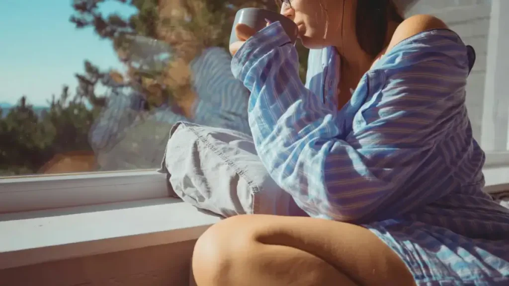 Dziewczyna z kubkiem kawy na parapecie patrzy przez okno – chwila spokoju i minimalizm w życiu.